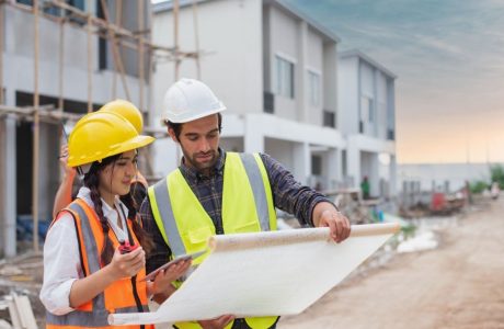 Caucasian males and Asian female builders, architector and engineers with draft plan of building and laptop computer talking on constructing site